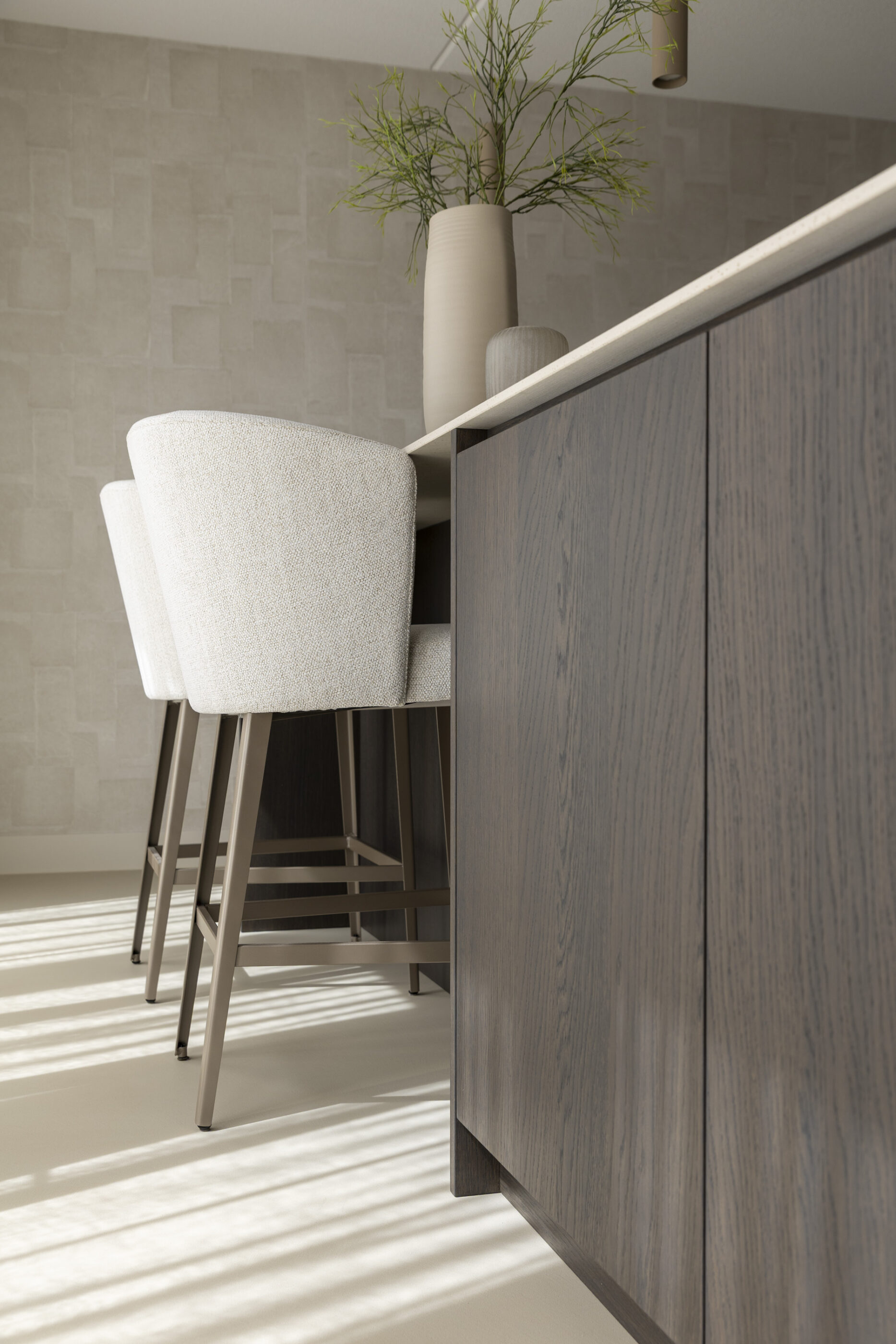 Detail van kookeiland in donker eikenfineer met beige barstoelen en een strak afgewerkt werkblad in lichte steenlook.